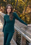 A woman with long brown hair wears the Woolx Hadley Ribbed Crew in Deep Timber, standing on a wooden bridge with her arm on the railing. Trees and water are in the background under a sunny sky.