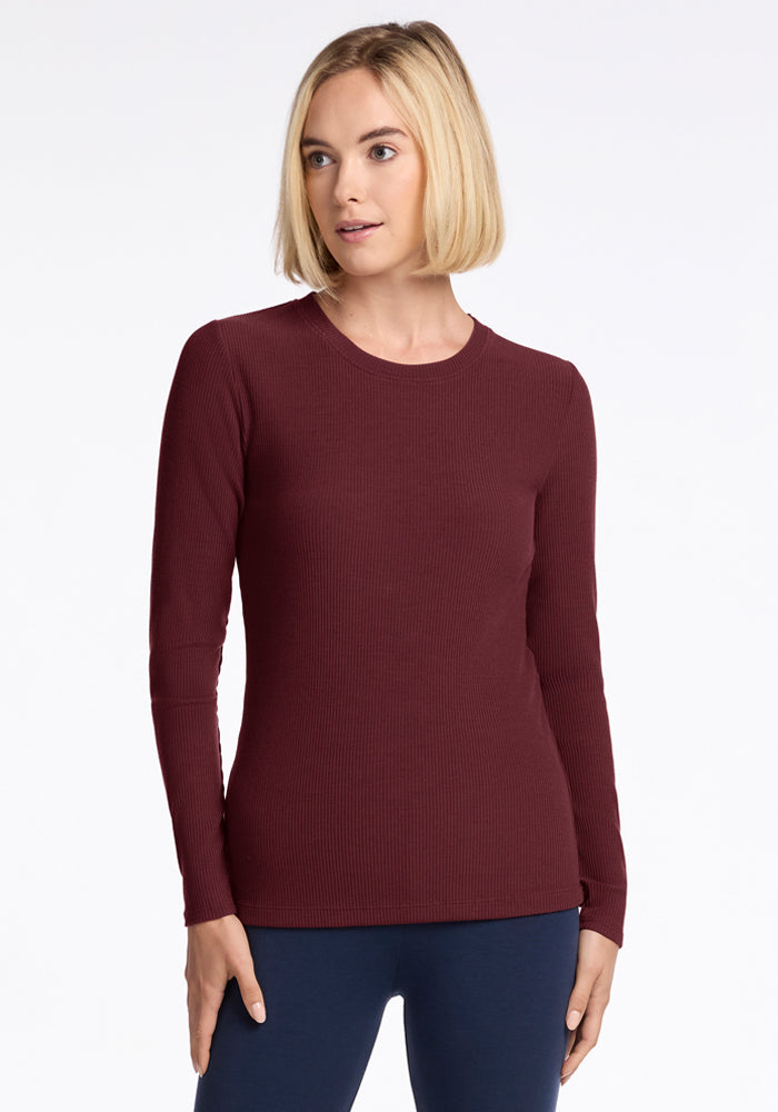 A woman with straight blonde hair wears the Woolx Hadley Ribbed Crew in Toasted Garnet, paired with navy pants—perfect travel attire—as she stands against a plain white background, gazing slightly to the side.