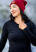 A woman in the Woolx Hannah Long Sleeve Top - Black and a red beanie stands outdoors, looking to the side with a slight smile.