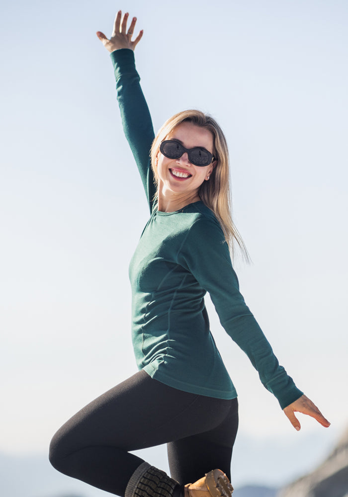 A smiling woman in sunglasses poses outdoors against a bright sky, wearing the Woolx Hannah Long Sleeve Top in Riverbank Green, black leggings, and boots with one arm raised and one leg bent.