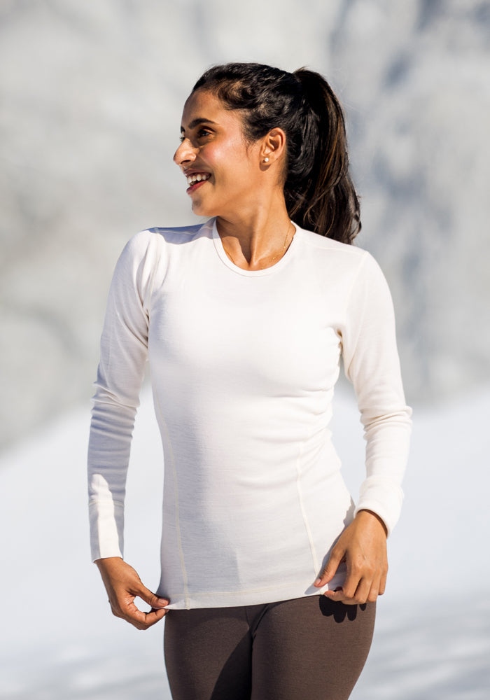A woman stands outdoors in the snow, smiling and looking to the side. She wears the Woolx Hannah Long Sleeve Top in Buttercream with brown leggings, her hair tied back in a ponytail.