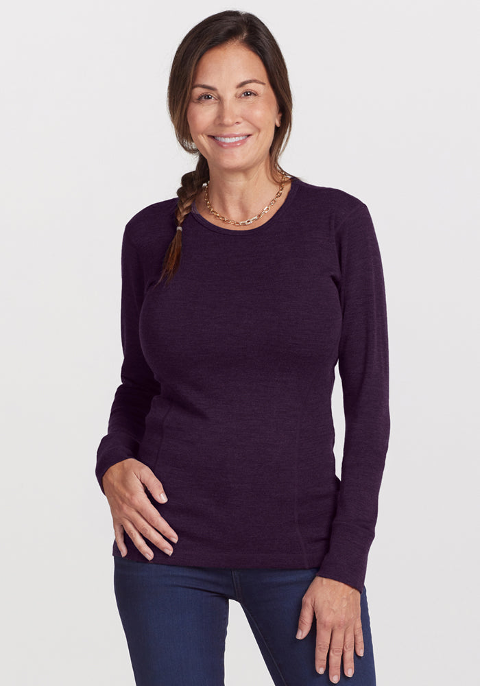 A woman with a side braid, wearing the Woolx Hannah Long Sleeve Top in Deep Plum and blue jeans, smiles at the camera against a plain light background.