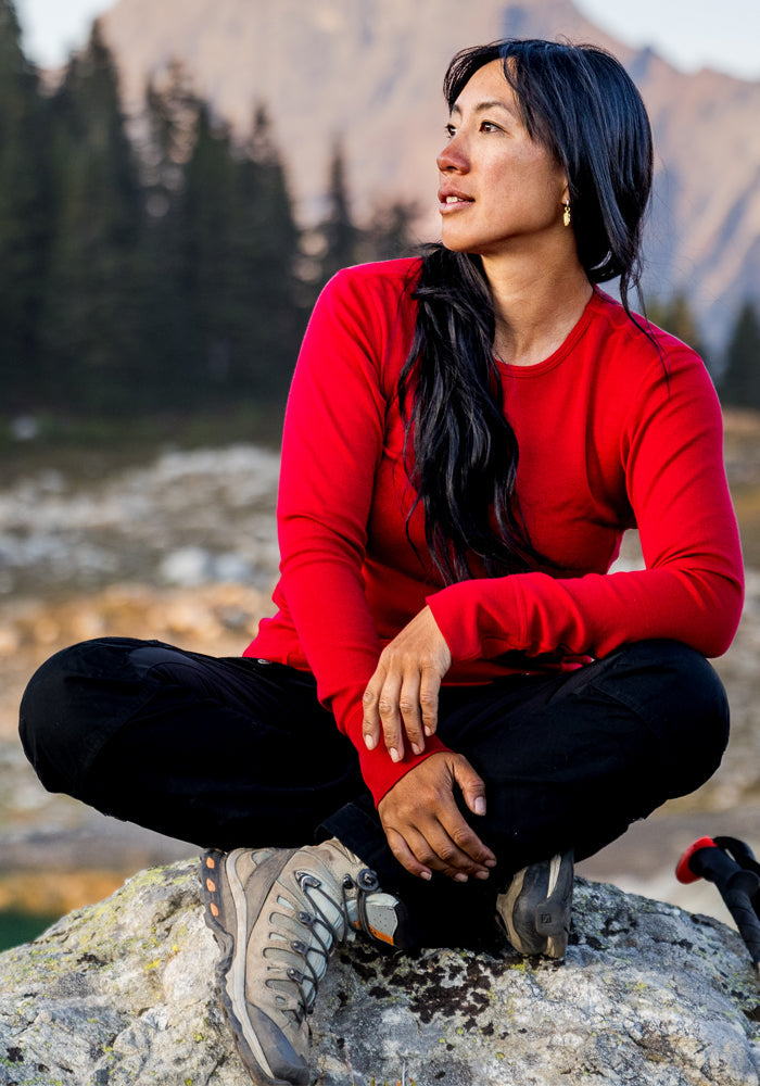 A woman in a Woolx Hannah Long Sleeve Top - Maraschino Cherry sits cross-legged on a rock, mountains and trees behind her.