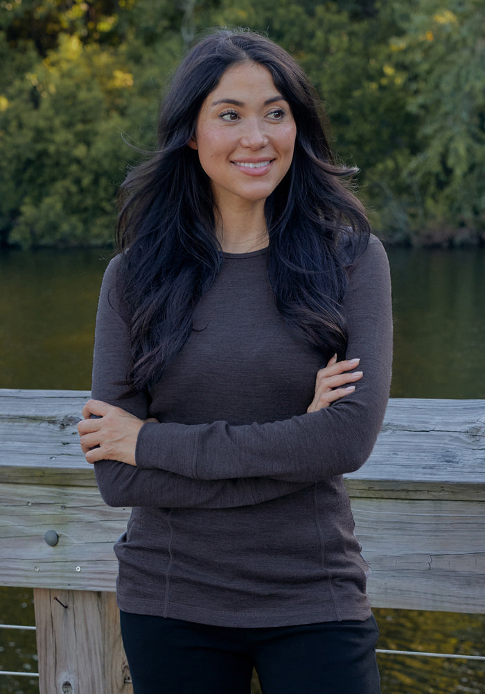 Woman with long dark hair, smiling outdoors in a Woolx Hannah Long Sleeve Top - Cinder Melange by a wooden railing.
