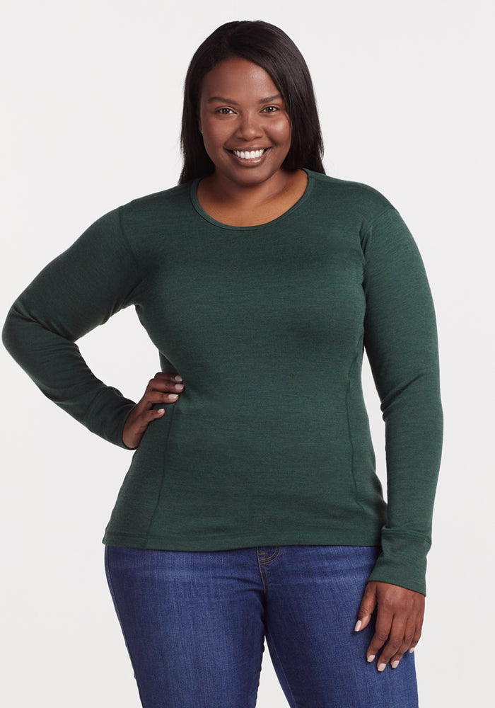 A woman wearing the Woolx Hannah Long Sleeve Top in Forest and blue jeans stands against a plain white background, smiling with one hand on her hip.