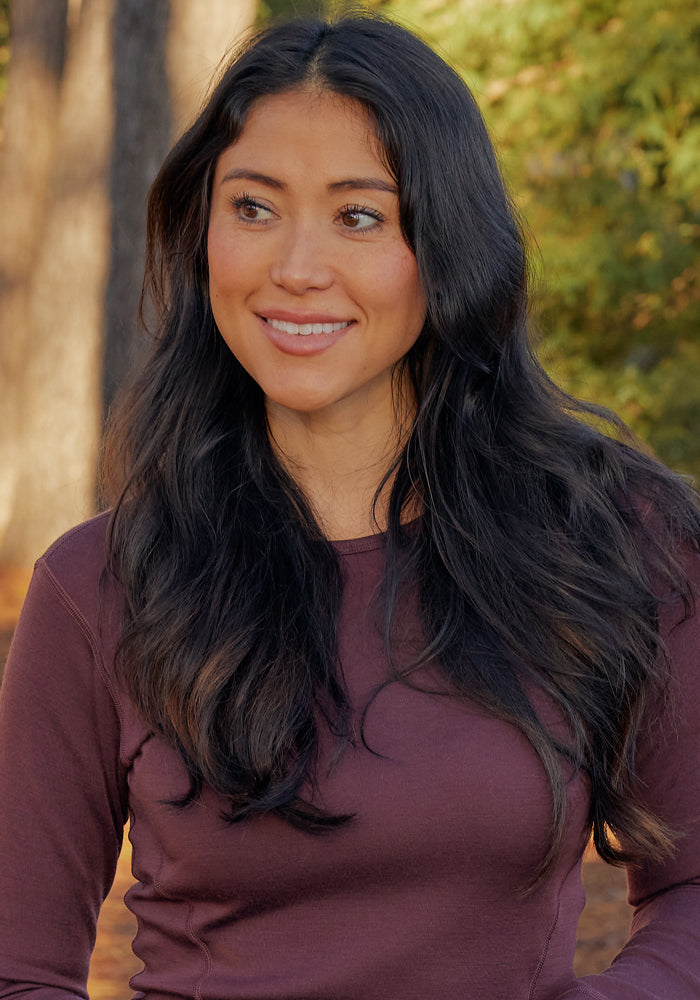 A woman with long dark hair stands outdoors among trees and sunlight, smiling softly and wearing the Woolx Hannah Long Sleeve Top in Chocolate Fig (Final Sale).