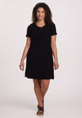 A curly-haired person wears the Harlowe Ribbed Dress by Woolx in black, paired with sandals, against a plain white background, smiling with hands relaxed by their sides. | Kisha is 5’8”, wearing a size M