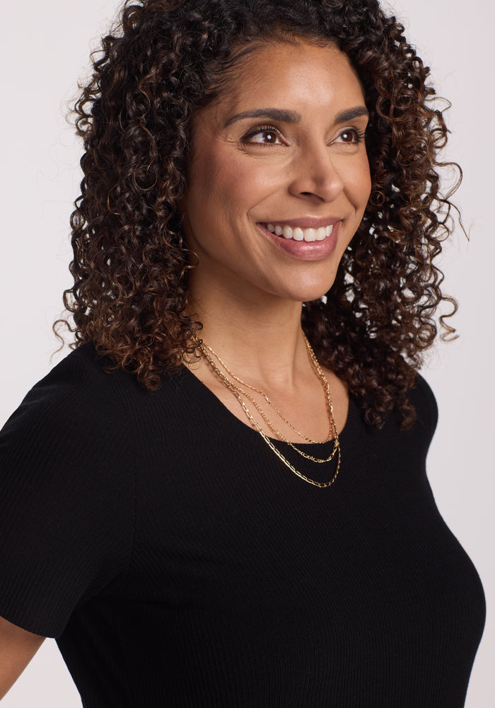 A smiling woman with curly hair, wearing a Woolx Harlowe Ribbed Dress in black and layered gold necklaces, looks to the side against a plain background.