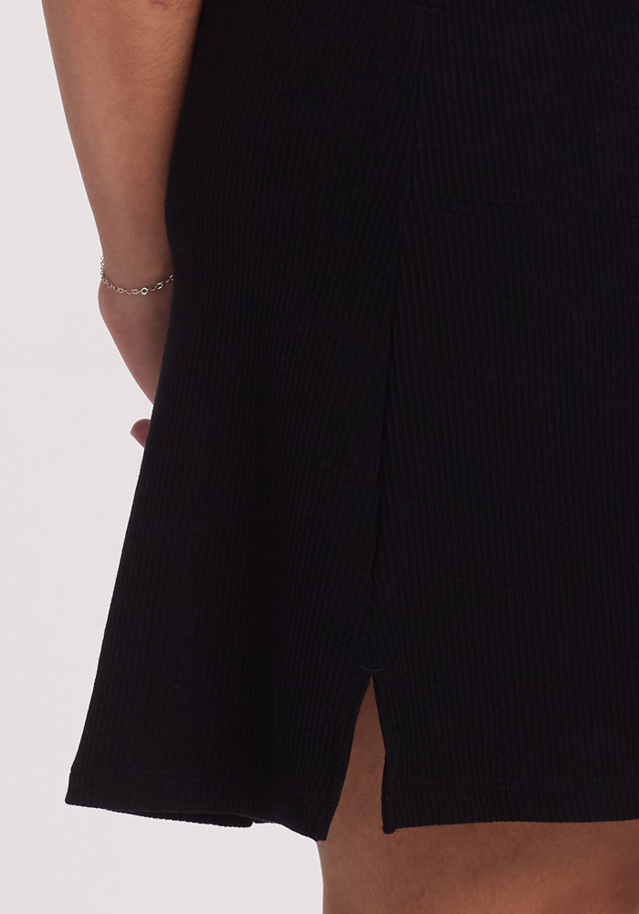 The person is wearing the Harlowe Ribbed Dress by Woolx in black, featuring a slit that reveals part of the leg. A delicate silver bracelet graces the wrist.