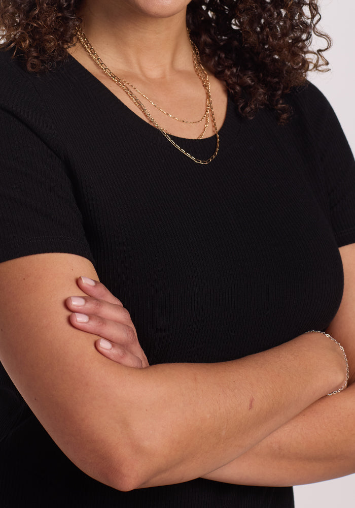 A person with curly hair wearing the Harlowe Ribbed Dress - Black from Woolx, accessorized with layered gold necklaces and crossing their arms. The image focuses on their shoulders and upper body.