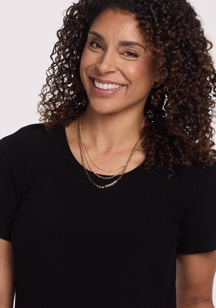 A smiling person with curly brown hair is wearing the Harlowe Ribbed Dress in black by Woolx, along with layered gold necklaces.