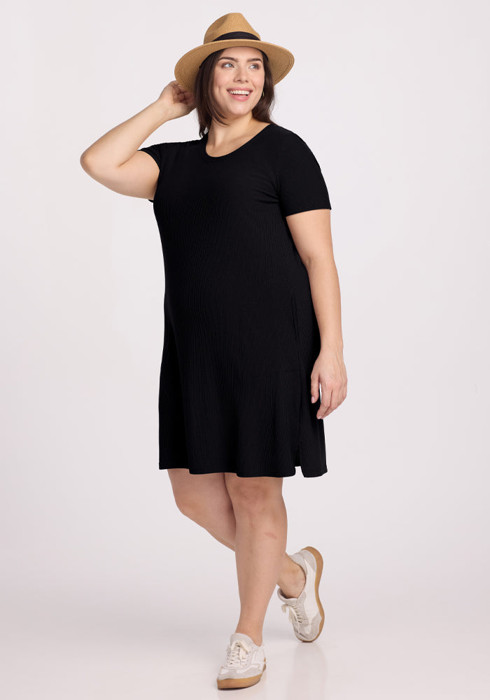 A person confidently poses in the Harlowe Ribbed Dress - Black by Woolx, paired with white sneakers. They smile with one hand on their straw hat against a plain white background, accentuating their casual and stylish look. | Juliana is 5'10", wearing a size XL