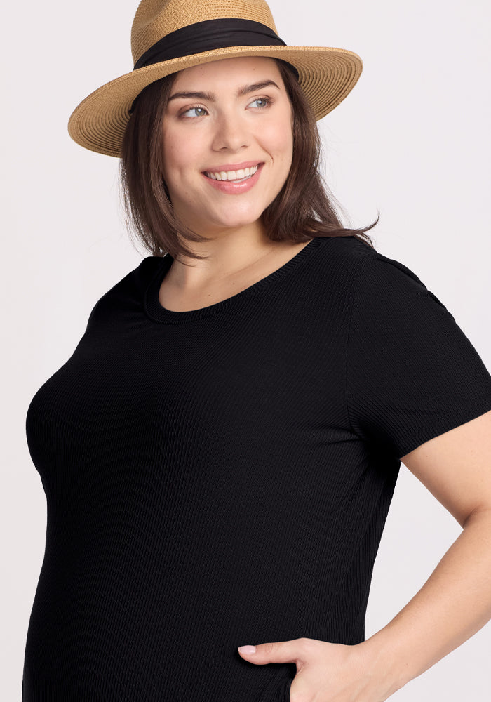A smiling person is wearing the Harlowe Ribbed Dress in black by Woolx along with a straw hat featuring a black band, all set against a plain, light-colored background.