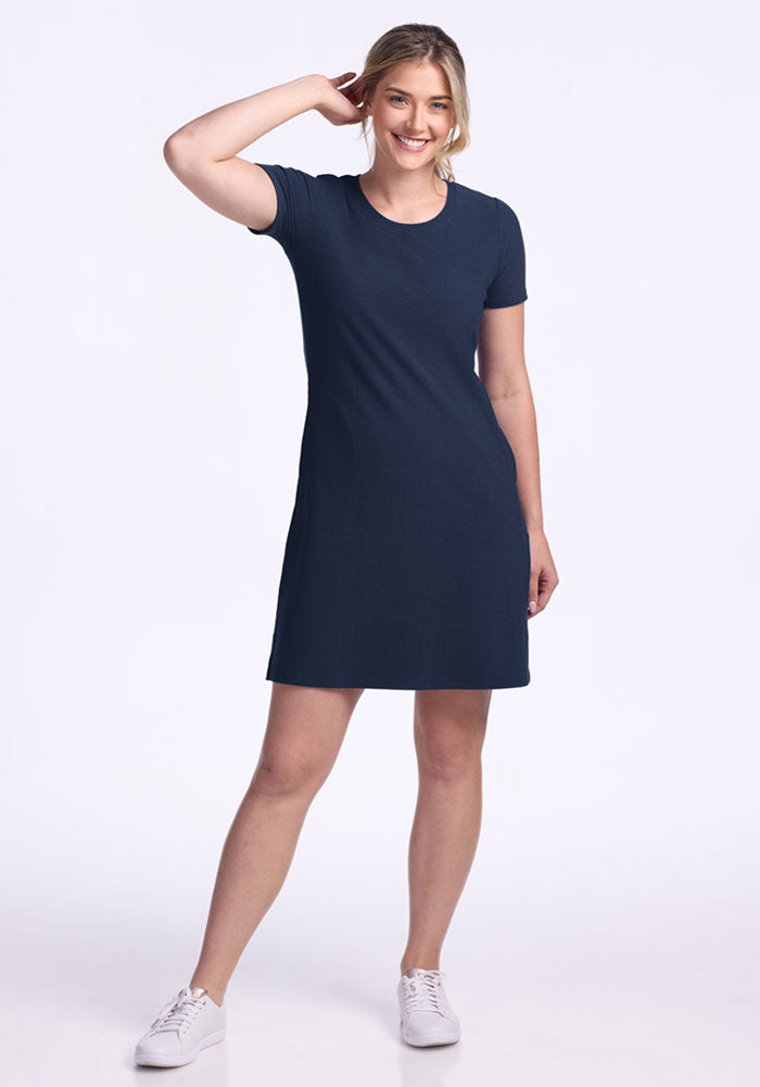 Woman smiling in a Woolx Harlowe Ribbed Dress in Deep Navy and white sneakers, standing against a white background.