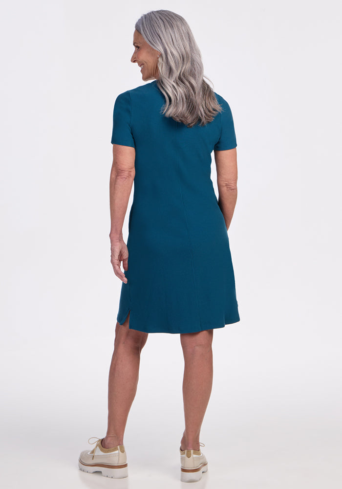 A woman models the Woolx Harlowe Ribbed Dress in Moroccan Blue, facing away, with beige shoes on a white background.
