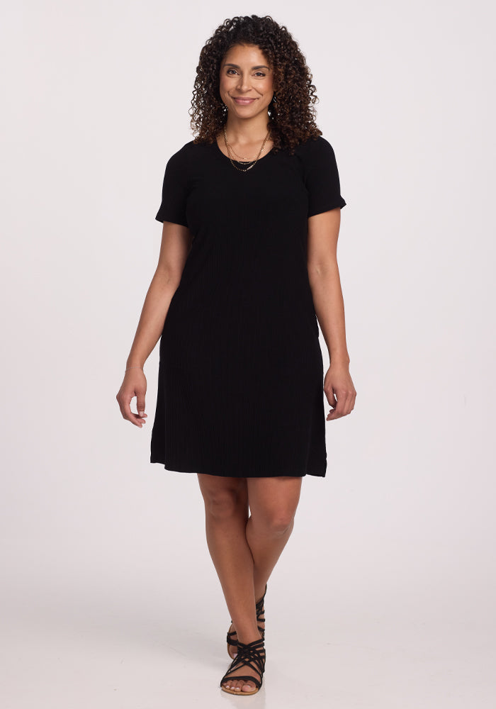 A curly-haired person wears the Harlowe Ribbed Dress by Woolx in black, paired with sandals, against a plain white background, smiling with hands relaxed by their sides. | Kisha is 5’8”, wearing a size M