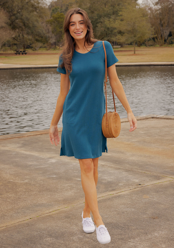 Woman in the Woolx Harlowe Ribbed Dress in Moroccan Blue, white sneakers, and round bag walking by a park lake.