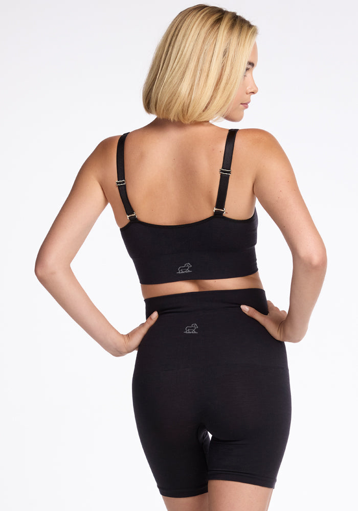 A person with blonde hair is standing with their back facing the camera, wearing a black Hayden Bra from Woolx and high-waisted shorts, offering comfortable support. Their hands are on their hips against a plain white background.