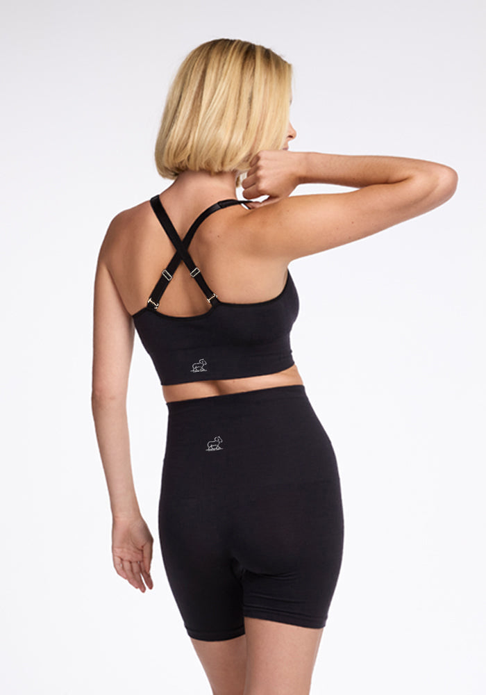 A woman with short blonde hair is facing away, wearing the Woolx Hayden Bra - Black made from luxurious merino wool and high-waisted black athletic shorts. The sports bra features an X-back strap design, offering comfortable support. The background is plain white.