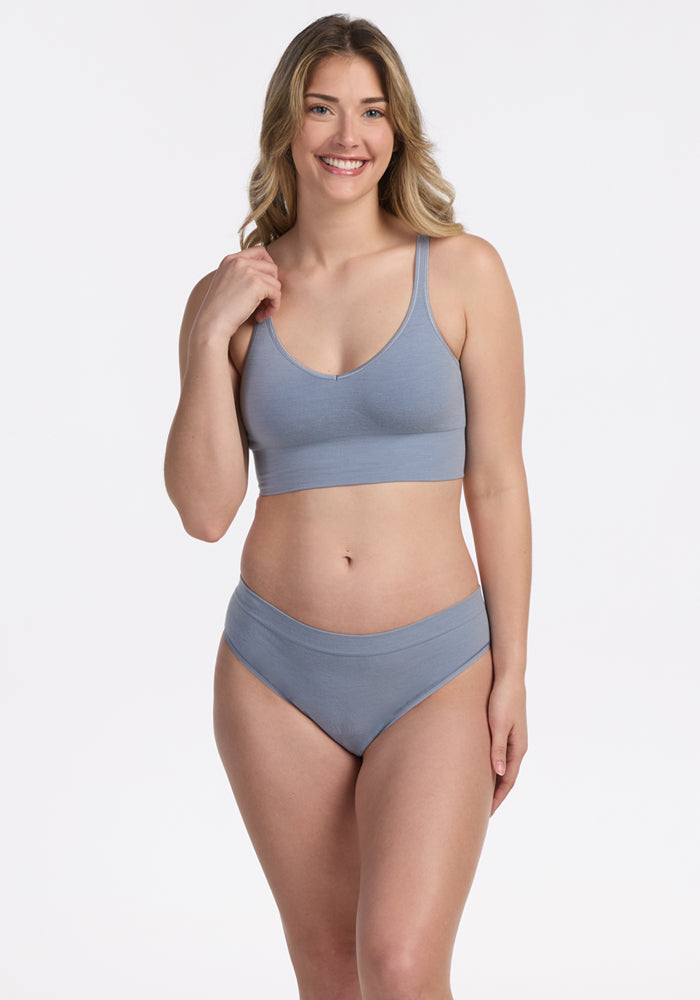 A woman with long, wavy hair smiles while modeling the Woolx Hayden Bra - Bluebell, a seamless comfort bra, with a matching underwear set against a plain white background.