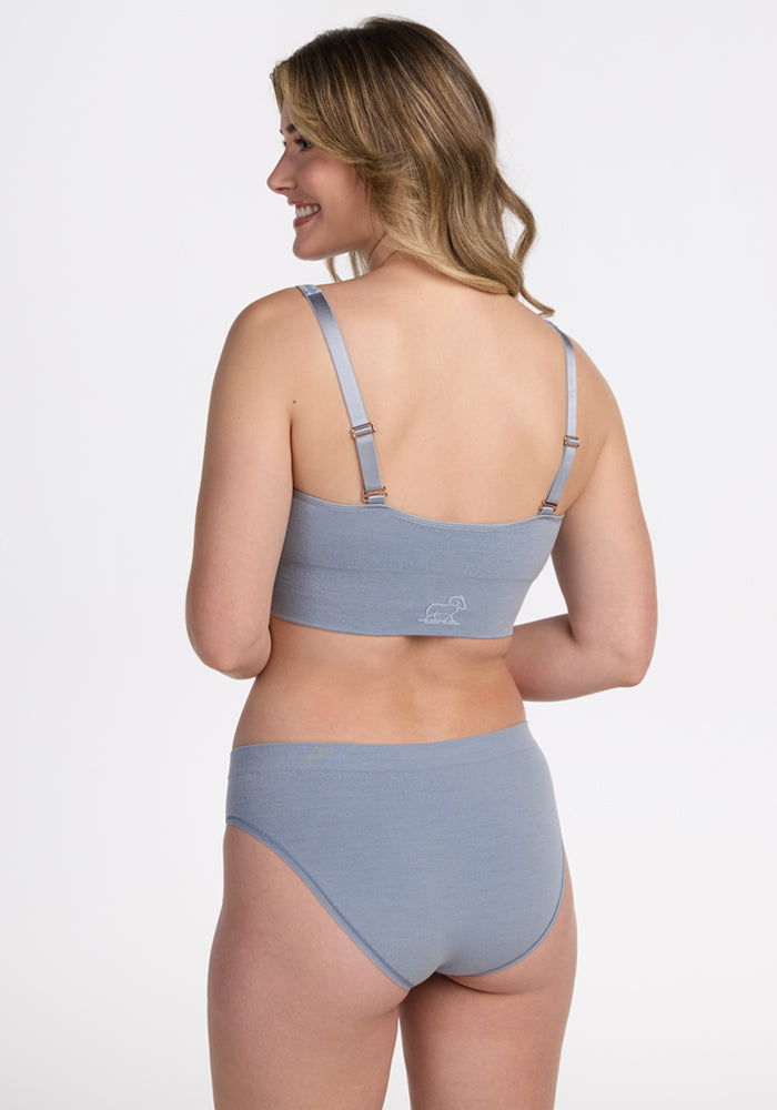 A woman with wavy, light brown hair smiles and stands sideways, modeling the Woolx Hayden Bra - Bluebell and matching underwear set against a plain white background.