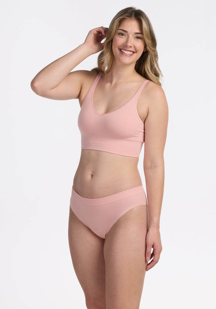 A woman with long, wavy blonde hair smiles while wearing the Woolx Hayden Bra in Rosewater with matching underwear, standing against a plain white background.