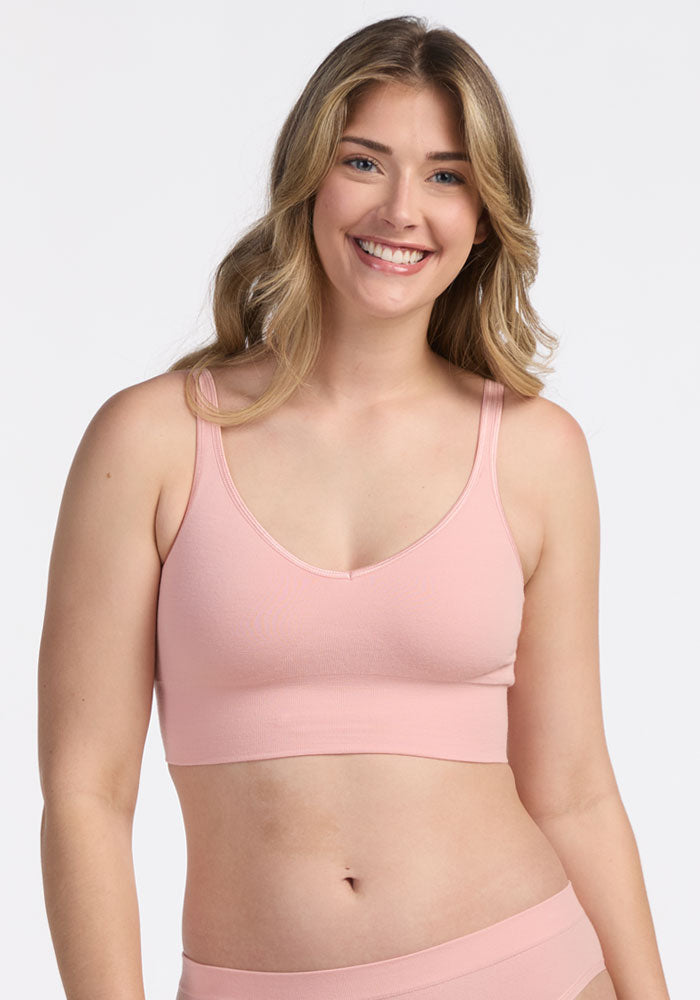 A woman with long, light brown hair smiles at the camera while wearing the Woolx Hayden Bra in Rosewater with matching bottoms. The background is plain white.
