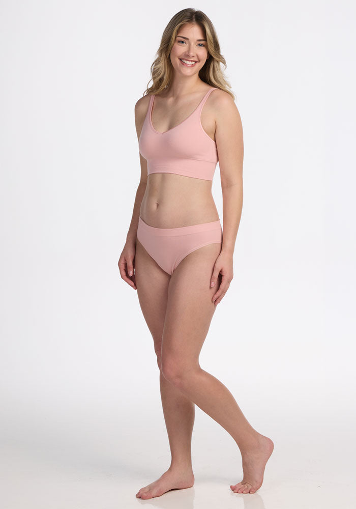 A woman with long, wavy hair smiles in a studio, wearing the Woolx Hayden Bra in Rosewater and matching underwear, posing naturally against a plain white background.