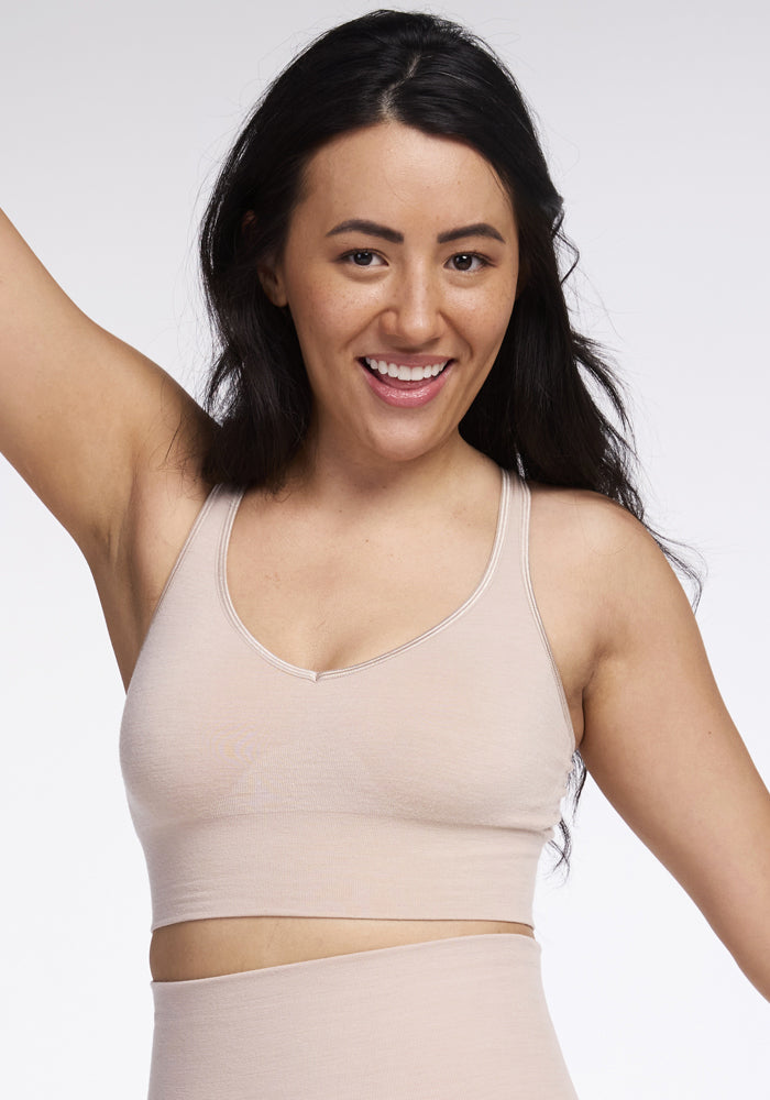 A person with long dark hair smiles and raises one arm, showcasing the seamless Hayden Bra - Sand by Woolx. They are posing against a plain white background, exuding confidence and elegance in their beige sportswear. | Sarah is 5'8", wearing a size S