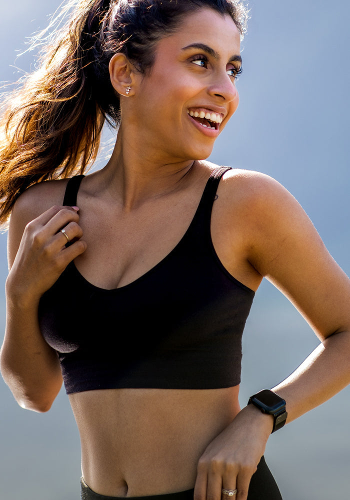 A smiling woman with long hair in a ponytail wears the Woolx Hayden Bra in Black and a smartwatch, standing outdoors in the sunlight.
