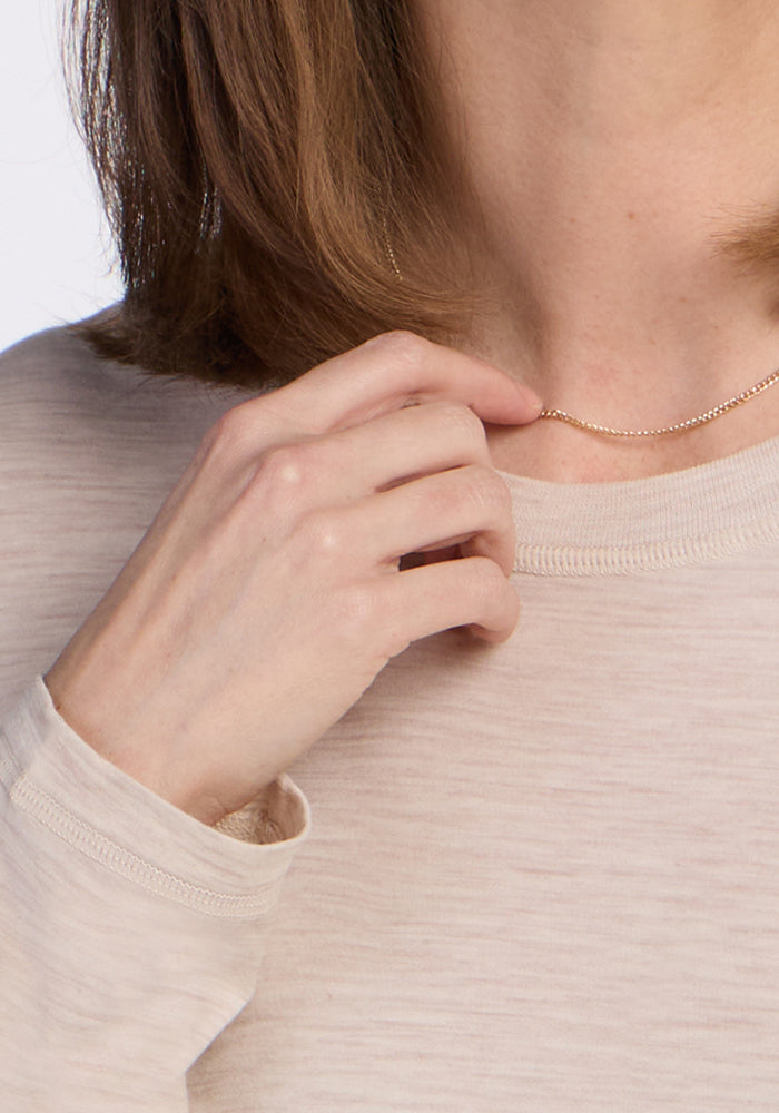 A person with light brown hair is wearing the Woolx Hazel Tunic in Cream Heather and holding a delicate gold chain necklace near their neck. The face is not visible.