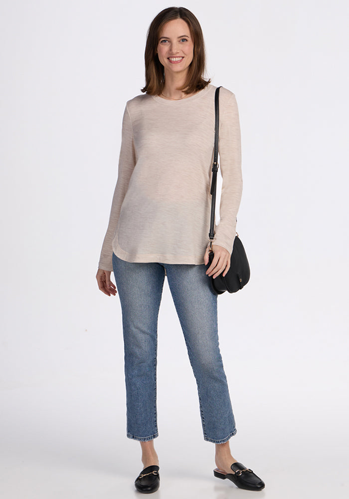 A smiling woman with shoulder-length brown hair wears the Woolx Hazel Tunic in Cream Heather, blue jeans, and black loafers. She carries a black crossbody bag and stands against a plain white background.