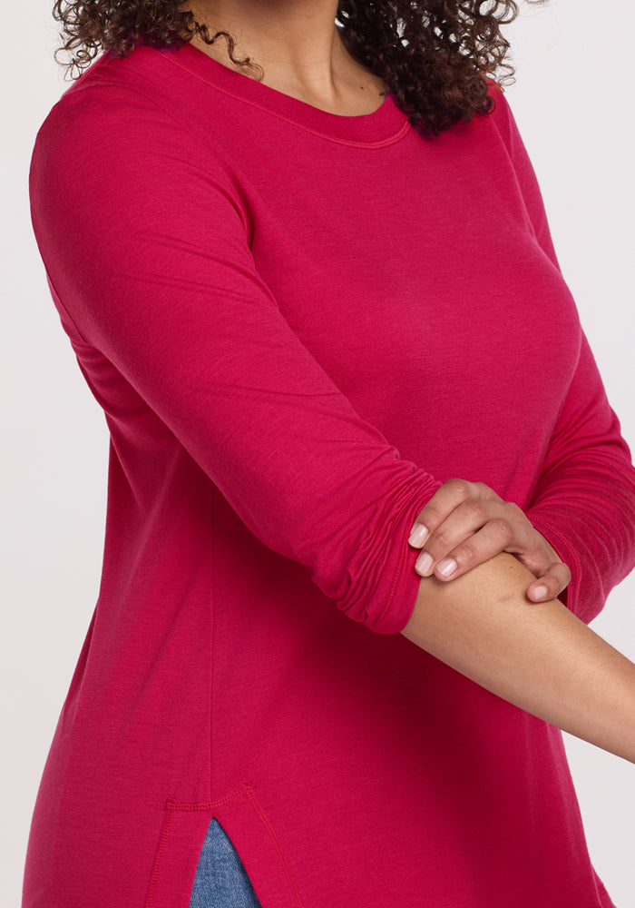 Woman adjusts sleeve of Woolx Hazel Tunic in Poppy Spark, shown shoulder to waist against a plain background.