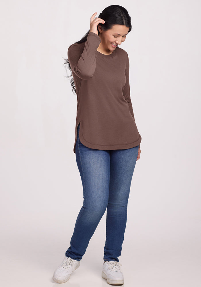 Woman in the Woolx Hazel Tunic - Peppercorn and blue jeans smiles, looking down, hand touching her hair.