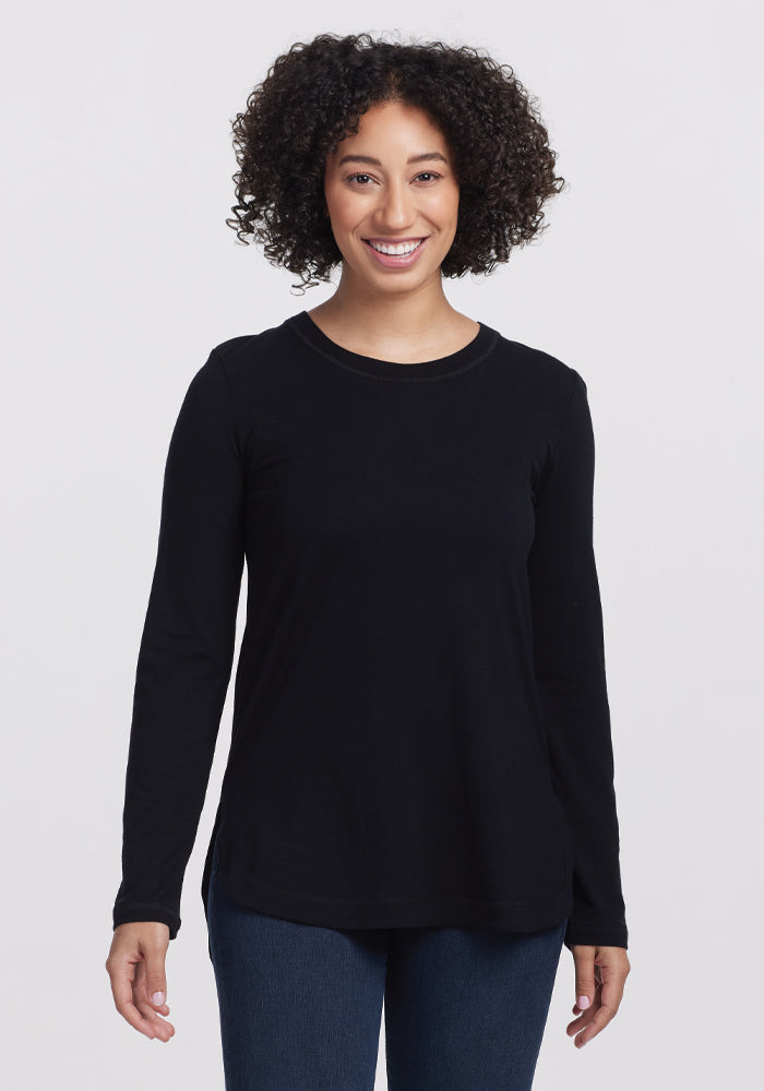 A person with curly hair smiles in the Woolx Hazel Tunic—an elegant long-sleeve black shirt made of temperature-regulating fabric. They pair it with jeans, standing against a plain white background. | Tori is 5'7", wearing a size S