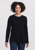A person with curly hair smiles in the Woolx Hazel Tunic—an elegant long-sleeve black shirt made of temperature-regulating fabric. They pair it with jeans, standing against a plain white background. | Tori is 5'7", wearing a size S