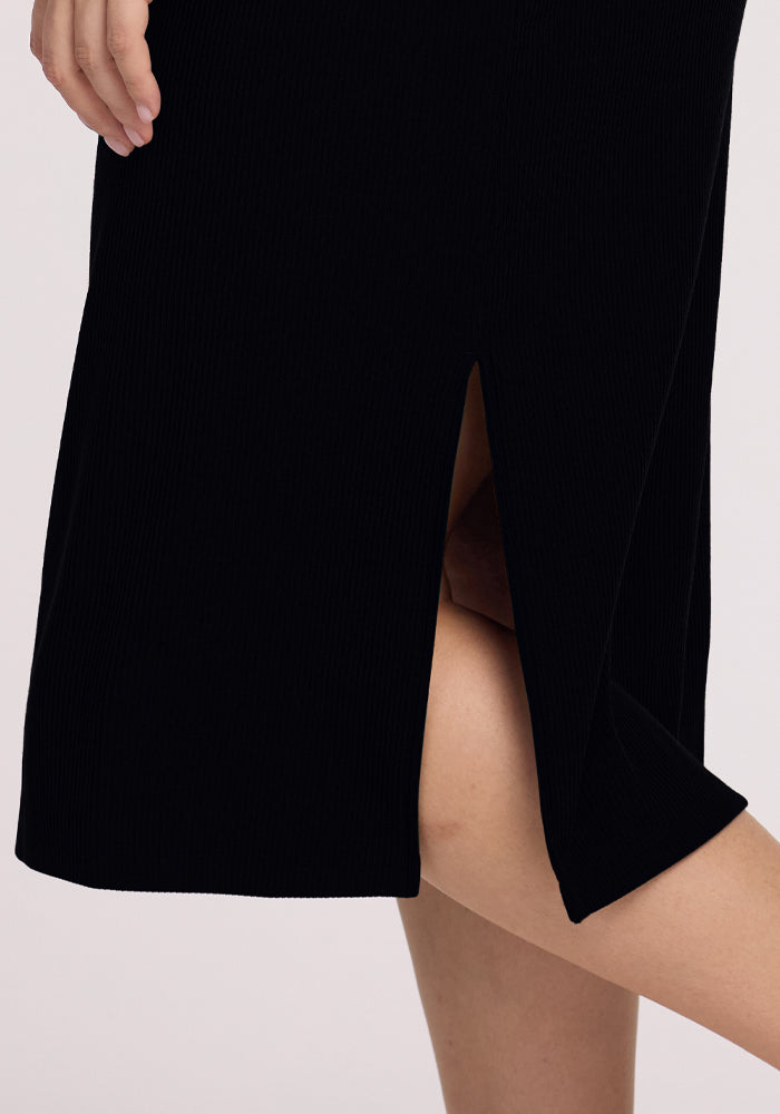 Close-up of someone wearing the Woolx Iris Ribbed Dress in black, showcasing its high side slit that reveals part of their leg. Their hand rests near the waist against a neutral background.