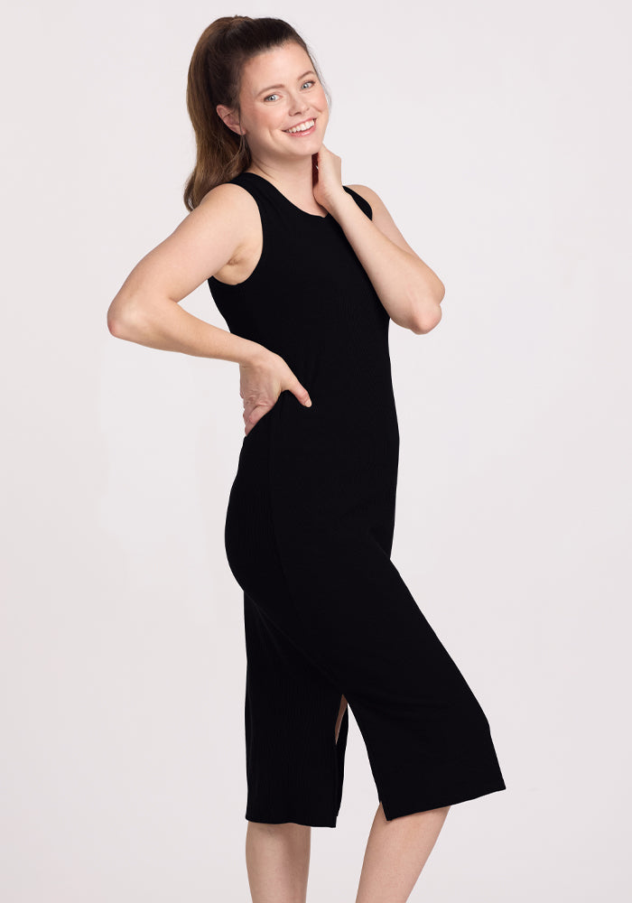 A person with long hair in a ponytail is smiling and posing sideways, wearing the sleeveless Iris Ribbed Dress in black with a side slit from Woolx against a plain background.