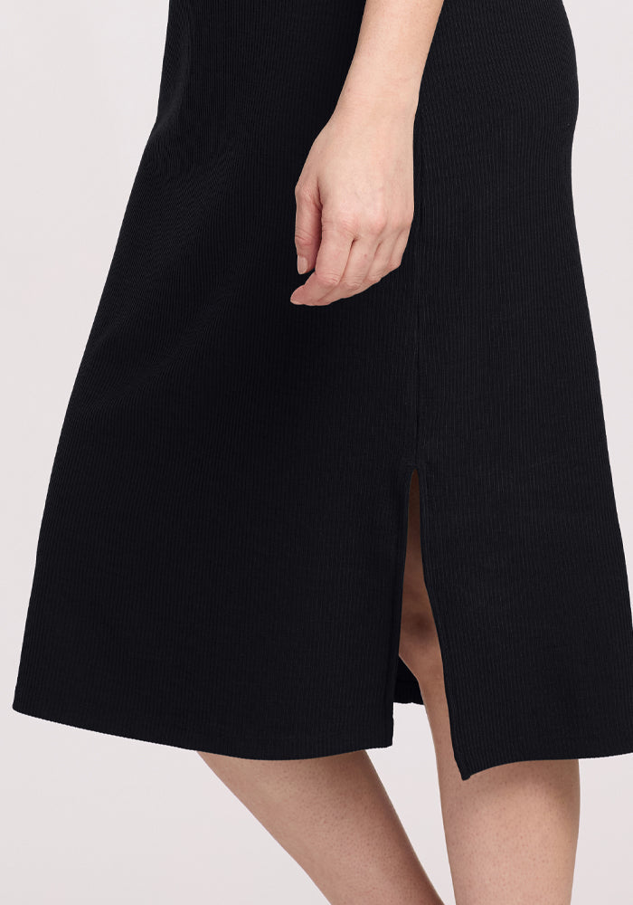 A person is wearing the Woolx Iris Ribbed Dress in black, featuring a side slit and knee-length hem. The individual's left hand is resting by their side against a neutral backdrop.