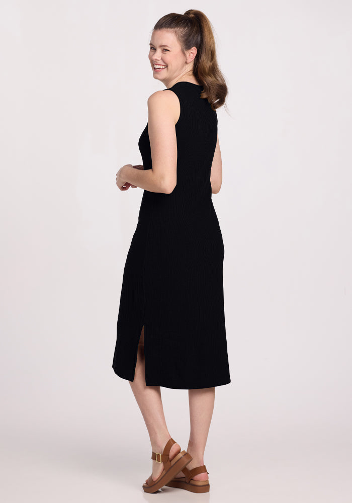 A person with long hair is smiling over their shoulder, wearing the Iris Ribbed Dress in black by Woolx and brown wedge sandals, standing against a plain light background.