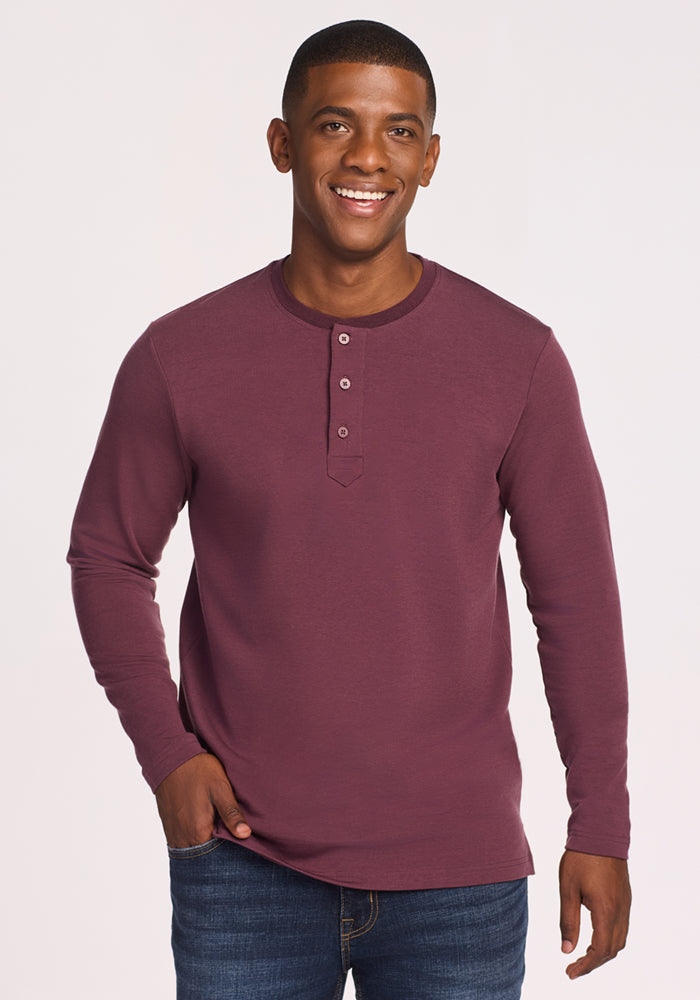 A smiling man wears the Woolx Jackson Henley in Grape Harvest with jeans, standing against a plain light background, one hand in his pocket.
