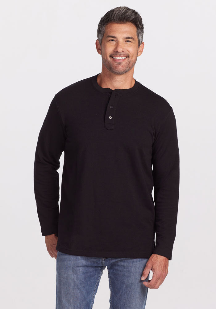 A man with short, dark graying hair and a beard is wearing the Jackson Henley - Black by Woolx, made of Merino wool, along with blue jeans. He is smiling and standing against a plain white background. | Matthew is 6', wearing a size L