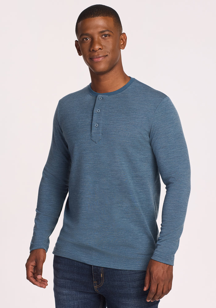 A man with short hair and a slight smile faces forward against a neutral light background, wearing the Woolx Jackson Henley • Final Sale - Steel Blue with dark jeans.