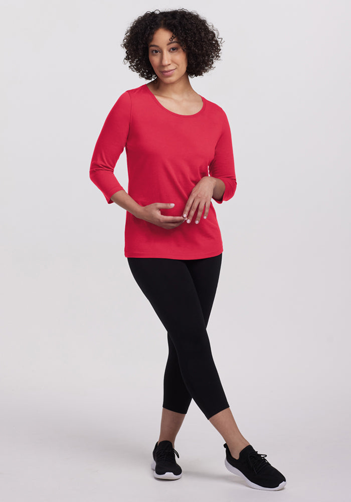 Woman with curly hair wears Woolx Jenny 3/4 Sleeve Crew Neck in Berry Ice Pop, black leggings, and sneakers, hands on stomach.