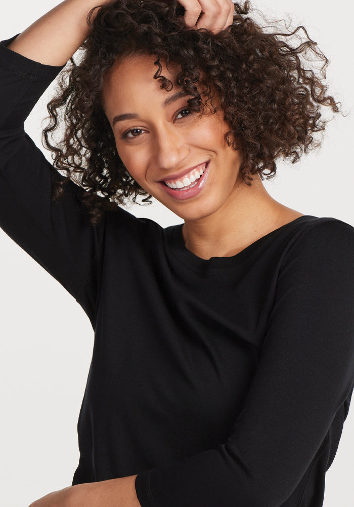 A person with curly hair smiles brightly while wearing the Jenny 3/4 Sleeve Crew Neck in black from Woolx, showcasing its classic crew neckline. They have one arm raised, touching their hair against a plain light background. This versatile piece adds understated elegance to any look.