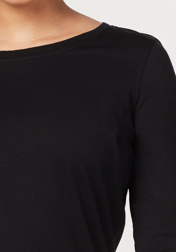 The image shows a close-up of someone wearing Woolx's Jenny 3/4 Sleeve Crew Neck in black. Crafted from merino wool, this staple features a classic crew neckline and offers a smooth and comfortable fit. It reveals part of the neck and shoulder against a plain background.