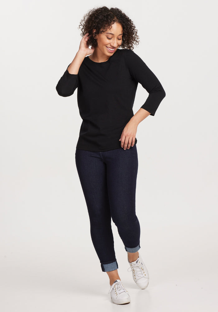 A person with curly hair smiles while standing in the Woolx Jenny 3/4 Sleeve Crew Neck in classic black, made of merino wool. They wear dark blue jeans with cuffs and white sneakers, posing against a plain, light background—a perfect addition to any versatile wardrobe.