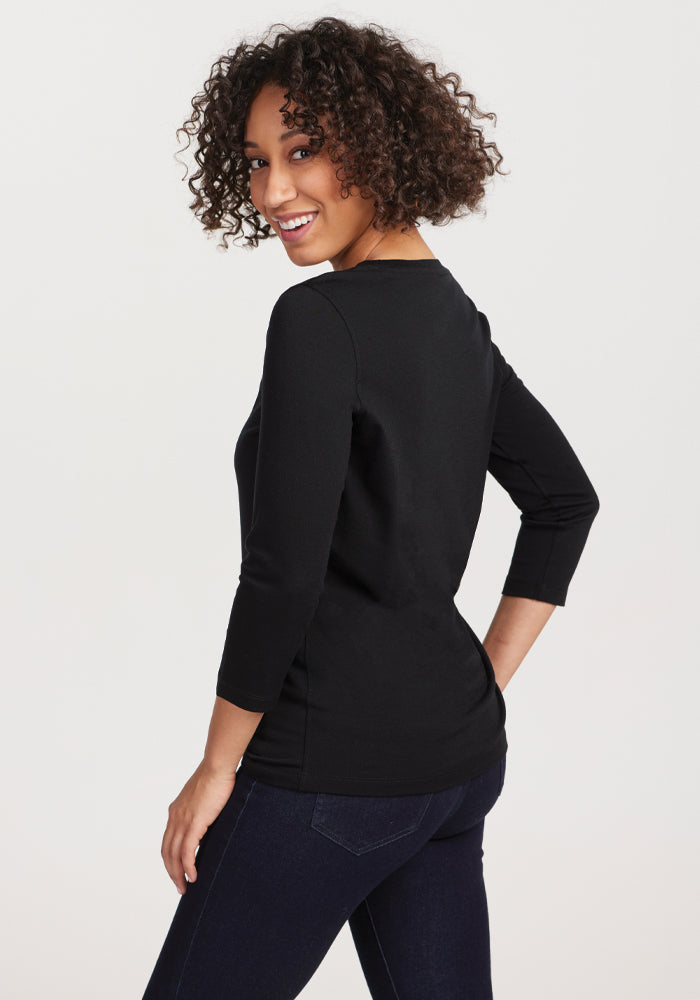 Against a plain white background, a curly-haired person smiles while looking over their shoulder. They are wearing the Jenny 3/4 Sleeve Crew Neck in black, made by Woolx from Merino wool, paired with dark jeans.