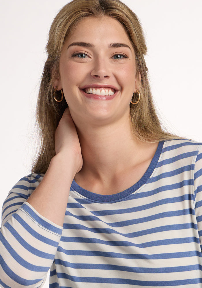 A smiling woman with long blonde hair wears the Woolx Jenny 3/4 Sleeve Crew Neck in Blue Sand Stripe, gold hoop earrings, and poses with one hand behind her neck against a plain white background.