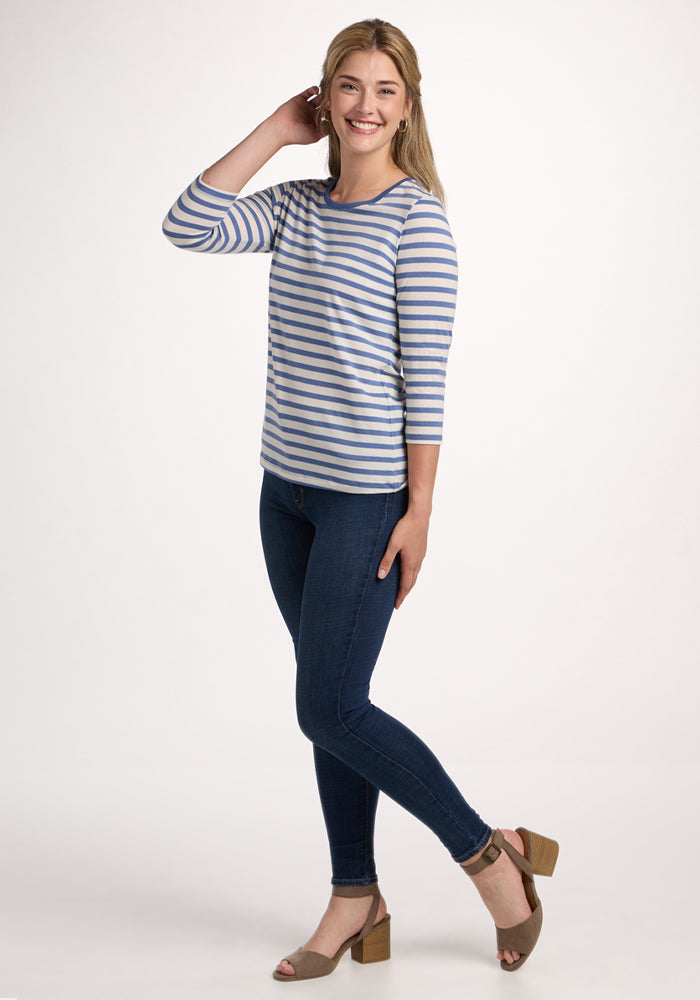 A woman smiles and poses in the Woolx Jenny 3/4 Sleeve Crew Neck in Blue Sand Stripe, paired with dark blue skinny jeans and tan heeled sandals, standing against a plain white background.